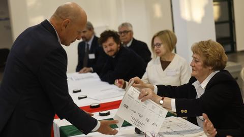 Ein Mann steht vor einem langen Tisch, an dem mehrere Menschen sitzen. Eine Wahlhelferin reicht ihm einen Wahlzettel.