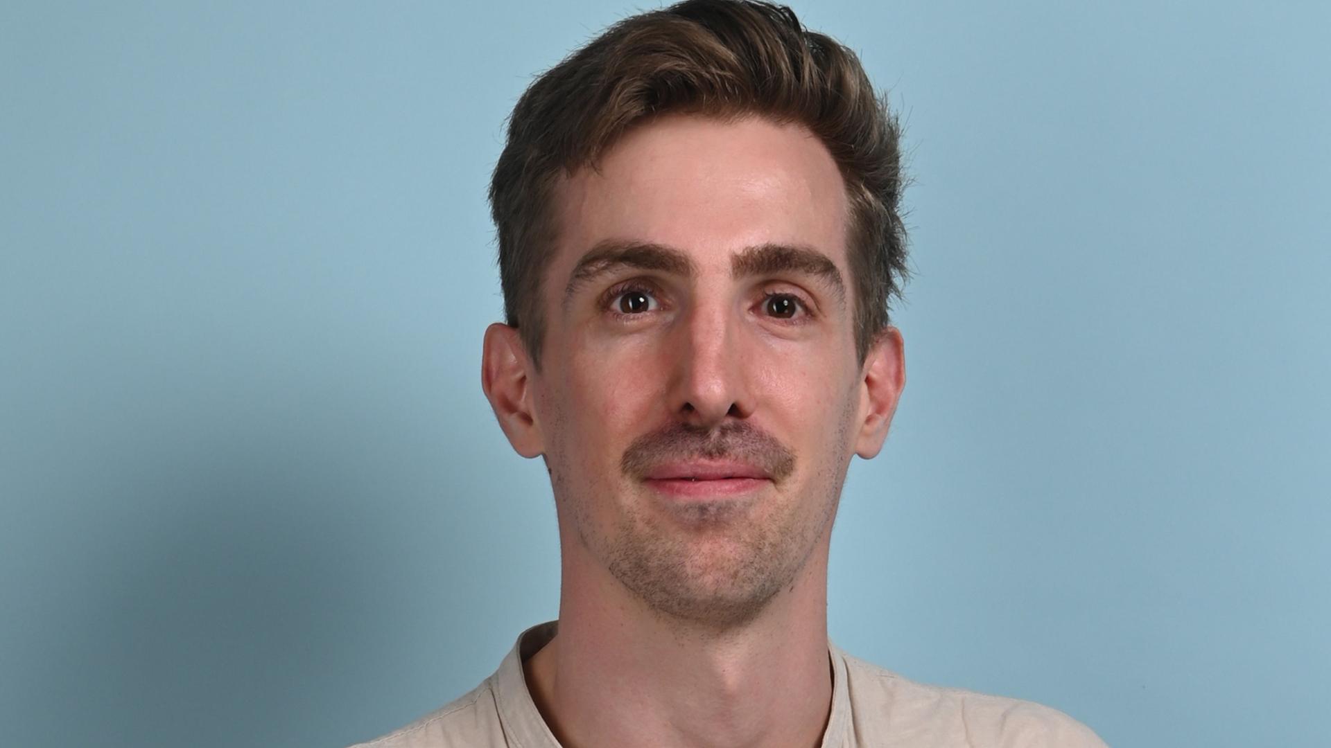 Portraitbild von Moritz Fehrle, der freundlich mit dunklen Haaren und Bart schaut, während er vor einer hellblauen Wand steht.