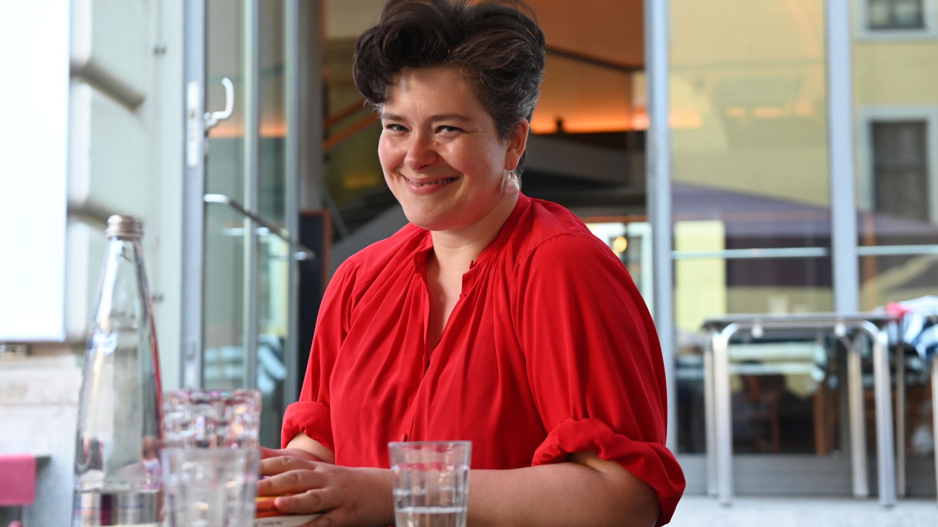 Nora Gomringer posiert lachend mit einem roten Oberteil an einem Tisch sitzend für ein Foto.