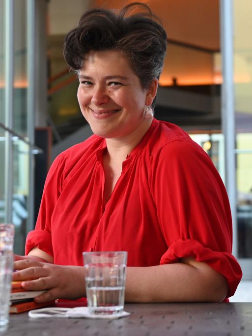 Nora Gomringer posiert lachend mit einem roten Oberteil an einem Tisch sitzend für ein Foto.