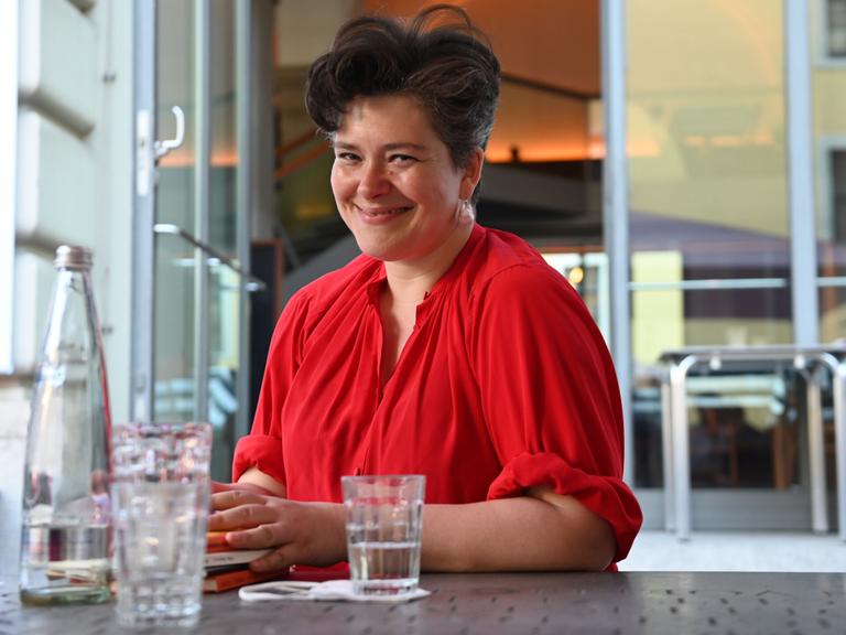 Nora Gomringer posiert lachend mit einem roten Oberteil an einem Tisch sitzend für ein Foto.