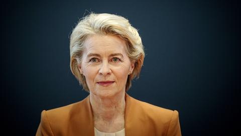 Ursula von der Leyen sitzt in einem senfgelben Sacko vor einer dunkelblauen Wand und schaut freundlich