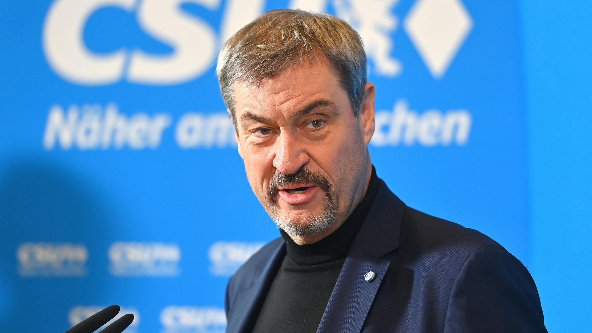 Mann mit kurzen grauen Haaren in einem blauen Jacket bei einer Rede vor einer blauen Wand, in der mit weißen Lettern CSU steht