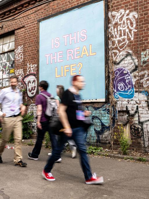 Verschwommene Menschen vor einem Plakat auf dem "Is This Real Life" steht.