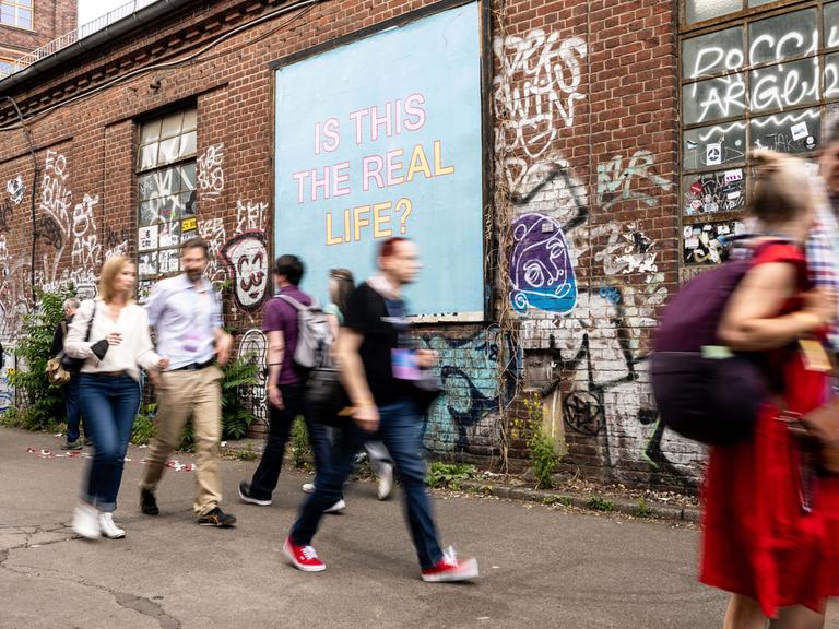 Verschwommene Menschen vor einem Plakat auf dem "Is This Real Life" steht.