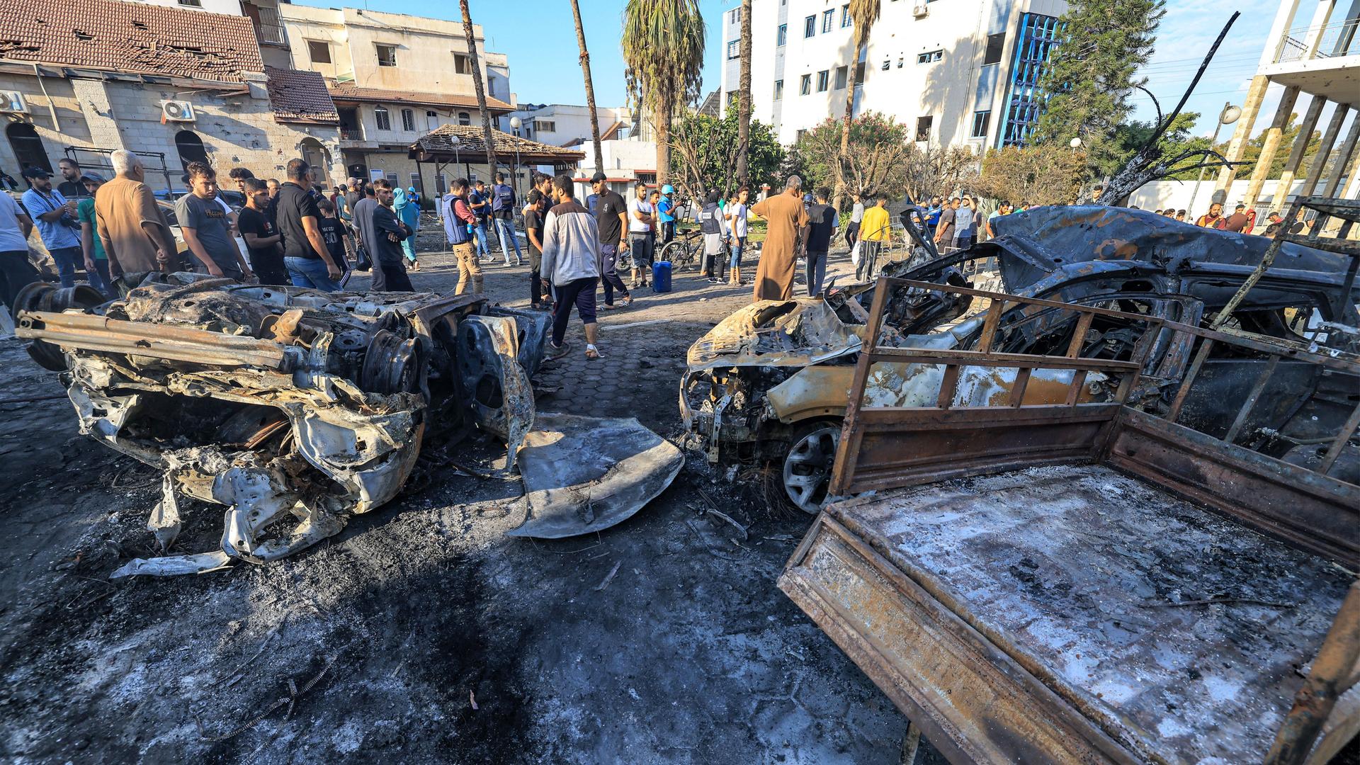 Menschen stehen auf einem Platz vor einem Kranken-Haus in Gaza-Stadt. Man sieht zerstörte Autos.

