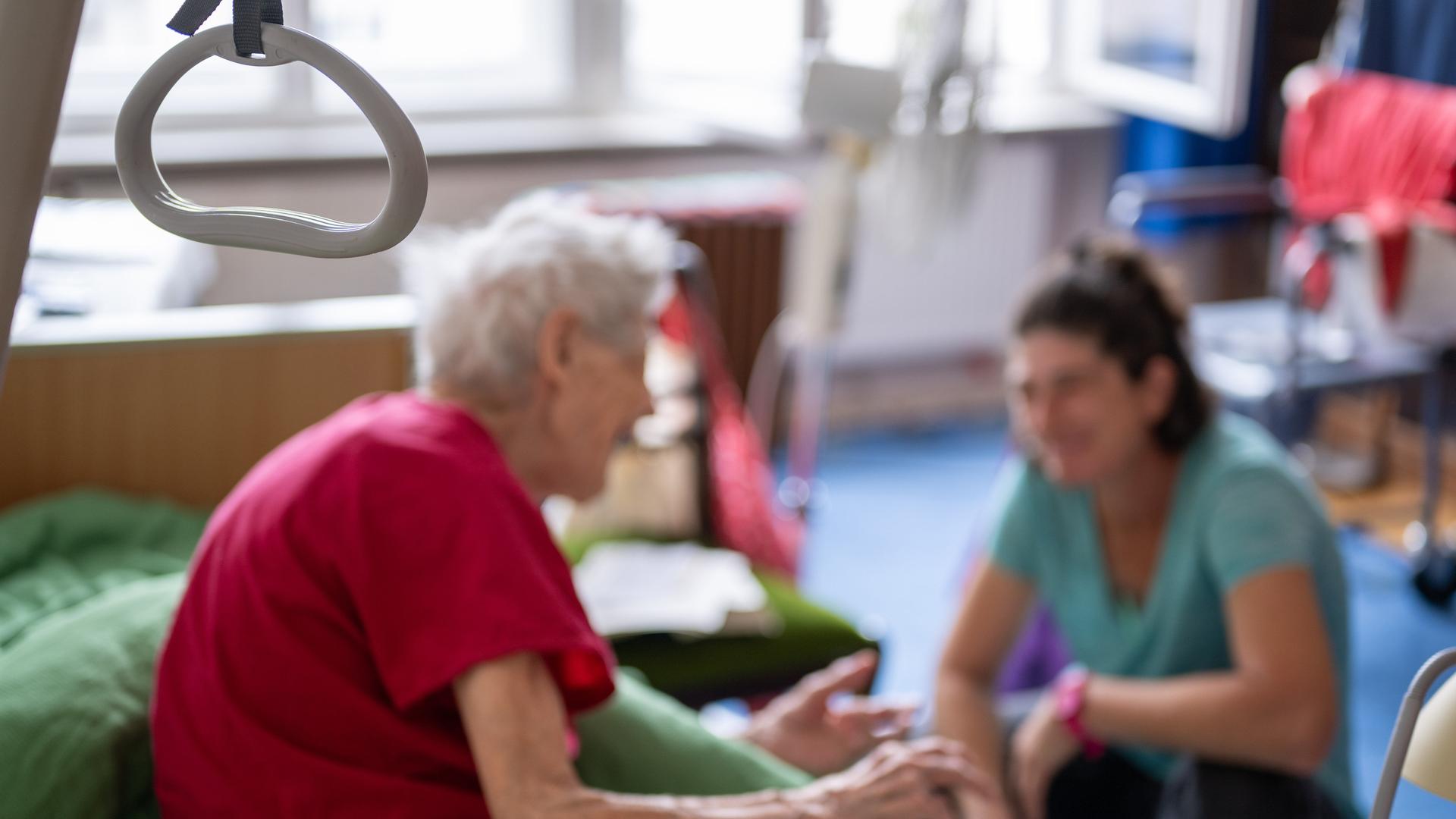 Eine Altenpflegerin hockt beim Hausbesuch vor dem Bett ihrer 97-jährigen Patientin und hört ihr zu.