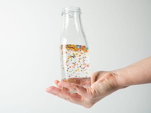 Nahaufnahme einer Hand, die eine mit Wasser gefüllte durchsichtige Glasflasche umschließt, in der winzige Mikroplastikpartikel schwimmen