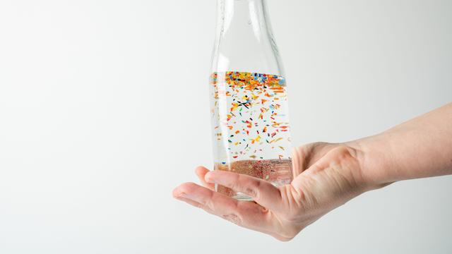 Nahaufnahme einer Hand, die eine mit Wasser gefüllte durchsichtige Glasflasche umschließt, in der winzige Mikroplastikpartikel schwimmen