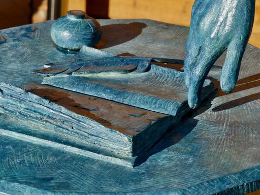 Das Detail einer Skulptur der Schriftstellerin Jane Austen zeigt eine Hand mit aufgeschlagenem Buch, Feder und Tintenfass. Das Detail einer Skulptur der Schriftstellerin Jane Austen zeigt eine Hand mit aufgeschlagenem Buch, Feder und Tintenfass.