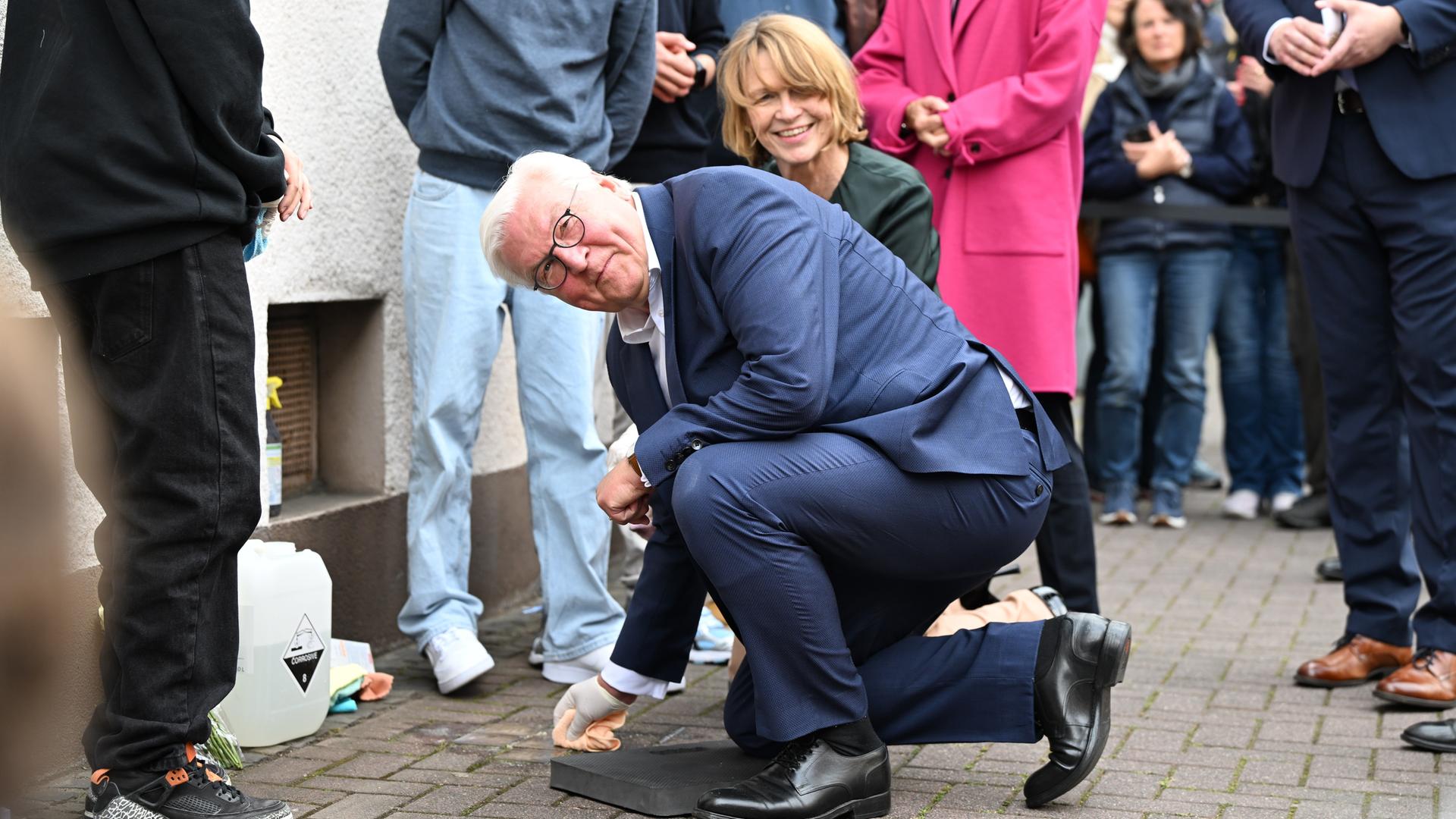 Steinmeier kniet am Boden und reinigt einen Stolperstein. Er trägt einen weißen Handschuh. Im Hintergrund ist Steinmeiers Frau, Elke Büdenbender, zu sehen.