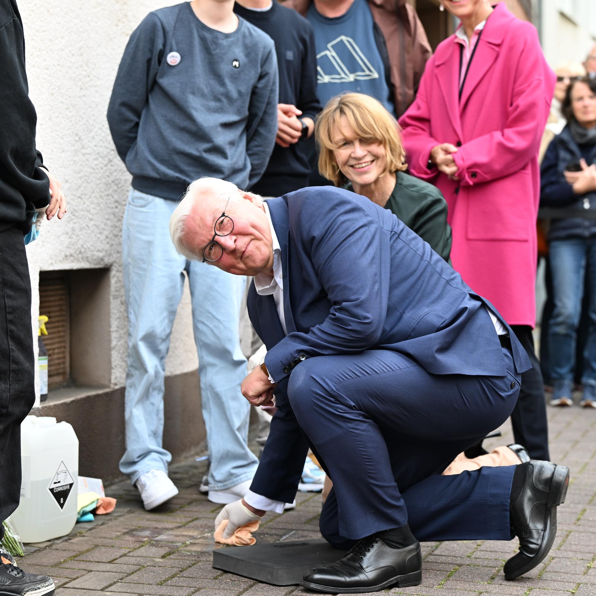 Steinmeier kniet am Boden und reinigt einen Stolperstein. Er trägt einen weißen Handschuh. Im Hintergrund ist Steinmeiers Frau, Elke Büdenbender, zu sehen.