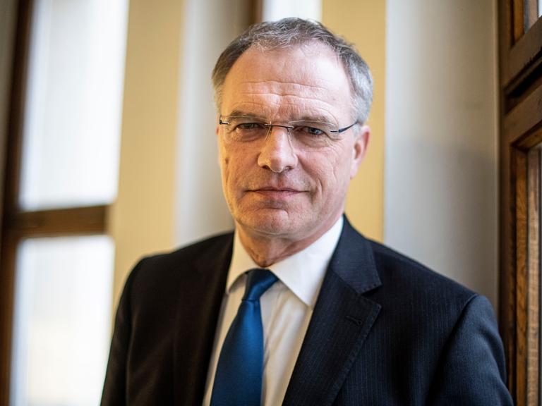 Stefan Raue schaut in die Kamera. Er trägt einen schwarzen Anzug mit dunkelblauer Krawatte und eine eckige, randlose Brille.