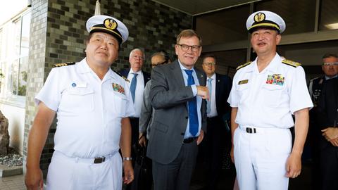 Johann Wadephul steht in einem grauen Anzug zwischen zwei unifomierten Angehörigen der japanischen Marine. Im Hintergrund sind weitere Männer in Anzügen zu sehen.
