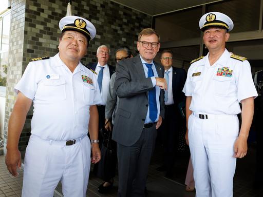 Johann Wadephul steht in einem grauen Anzug zwischen zwei unifomierten Angehörigen der japanischen Marine. Im Hintergrund sind weitere Männer in Anzügen zu sehen.