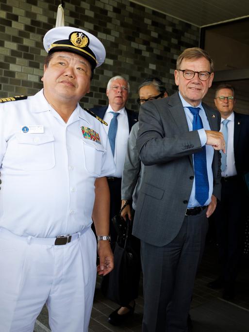 Johann Wadephul steht in einem grauen Anzug zwischen zwei unifomierten Angehörigen der japanischen Marine. Im Hintergrund sind weitere Männer in Anzügen zu sehen.