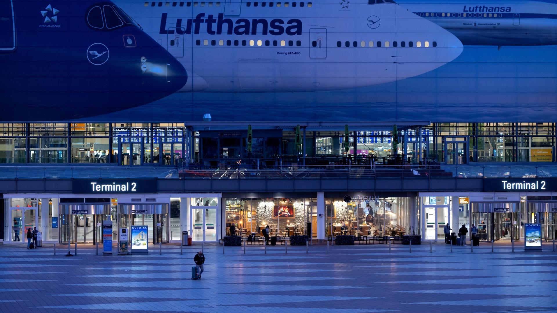 München: Passagiere gehen am Münchner Flughafen vor dem Terminal 2 an einem Plakat mit Flugzeugen der Lufthansa entlang.