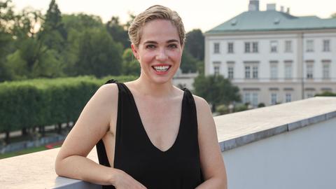 Verena Altenberger steht auf einem Balkon und lächelt in die Kamera. Sie trägt kurzes, blondes Haar und ein schwarzes ärmelloses Top.