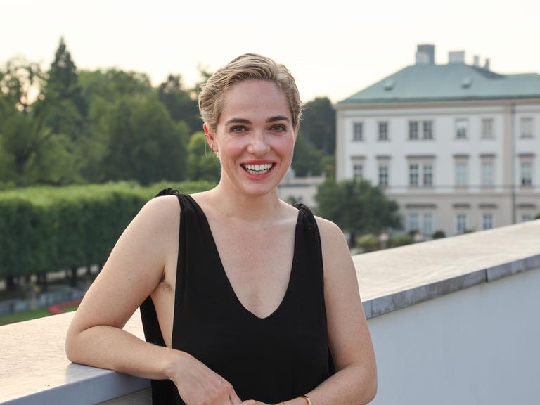 Verena Altenberger steht auf einem Balkon und lächelt in die Kamera. Sie trägt kurzes, blondes Haar und ein schwarzes ärmelloses Top.