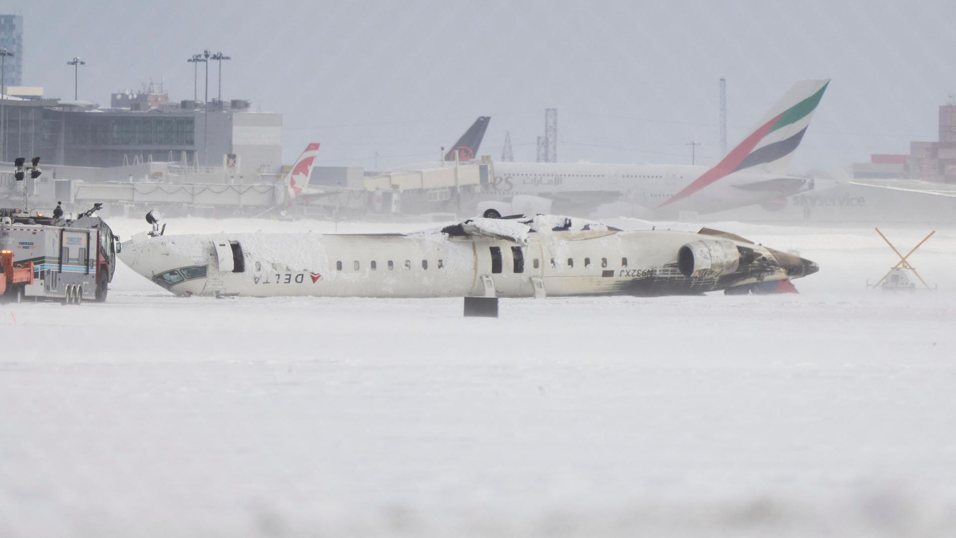 Das Bild zeigt ein Flugzeug der Delta Airlines auf dem Flughafen von Toronto, das nach der Landung kopfüber auf der Landebahn liegt. 