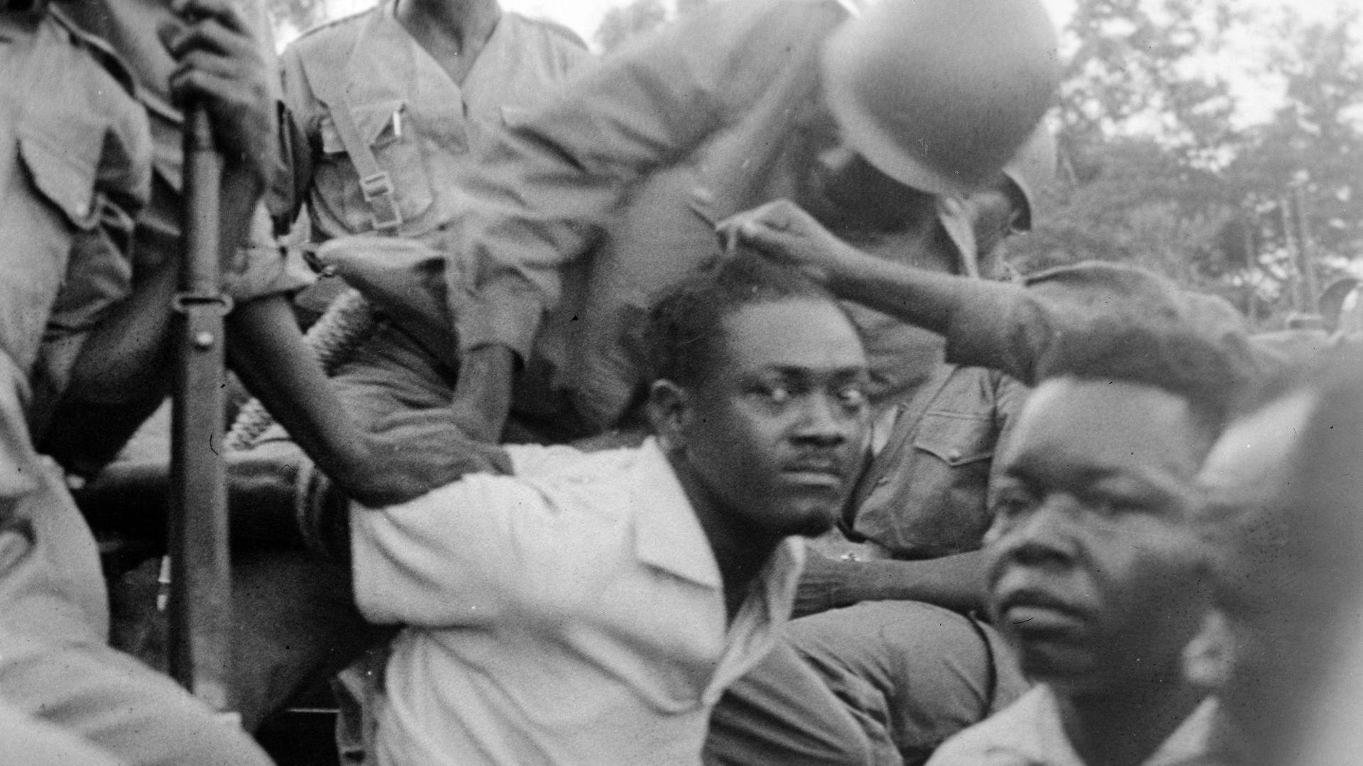 Historisches Schwarzweißfoto des von Soldaten festgenommenen Patrice Lumumba, der mit gehetztem Blick in die Kamera schaut. Historisches Schwarzweißfoto des von Soldaten festgenommenen Patrice Lumumba, der mit gehetztem Blick in die Kamera schaut.