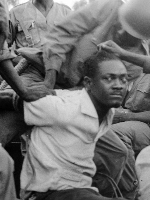 Historisches Schwarzweißfoto des von Soldaten festgenommenen Patrice Lumumba, der mit gehetztem Blick in die Kamera schaut.