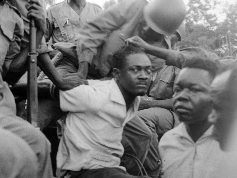 Historisches Schwarzweißfoto des von Soldaten festgenommenen Patrice Lumumba, der mit gehetztem Blick in die Kamera schaut.