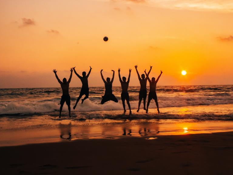 Eine Gruppe von Menschen springt am Strand vor einem Sonnenuntergang in die Luft sodass sich die Sihouetten der Personen klar abzeichnen.