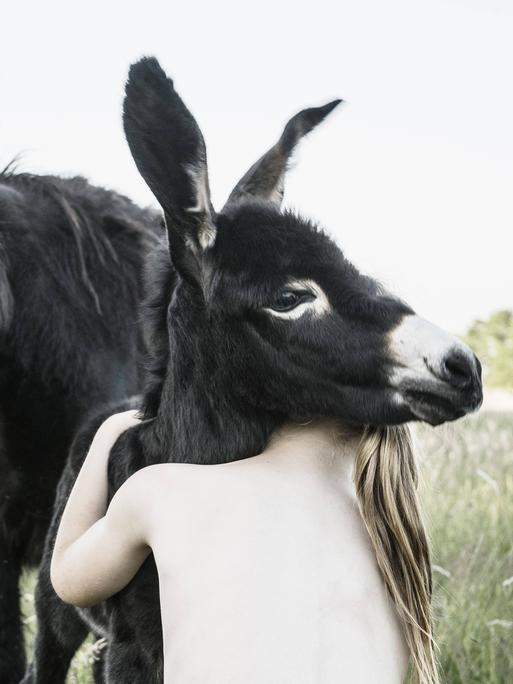 Ein Mädchen umarmt einen Esel, der nackte Oberkörper ist von hinten zu sehen, der Kopf versteckt sich hinter dem Esel.