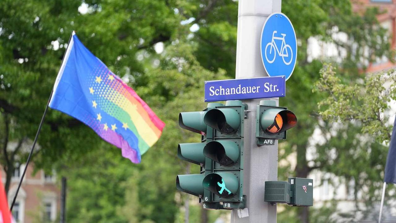 Das Bild zeigt eine Ampelanlage an einem Betonmast, an dem auch das Straßenschild "Schandauer Straße" hängt. Links davon ist eine Fahne zu sehen, auf der das Sternen-Motiv der EU übergeht in die Regenbogenfarben.