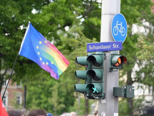 Das Bild zeigt eine Ampelanlage an einem Betonmast, an dem auch das Straßenschild "Schandauer Straße" hängt. Links davon ist eine Fahne zu sehen, auf der das Sternen-Motiv der EU übergeht in die Regenbogenfarben.