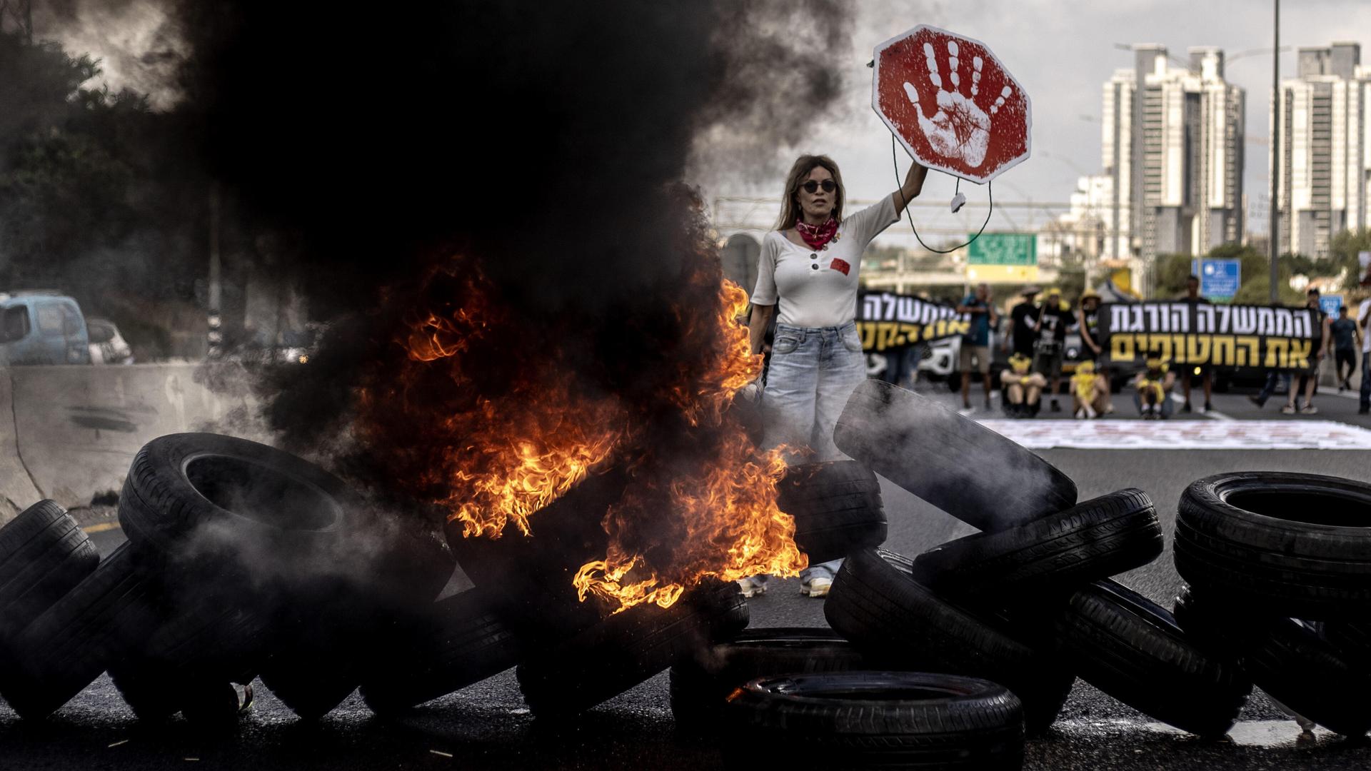 Demonstrantin in Tel Aviv (Israel) hält ein rotes Schild mit weißem Handabdruck in die Höhe; vor ihr liegt eine Barrikade aus brennenden Reifen, neben ihr steigt eine schwarze Rauchwolke in die Höhe (26.08.2025)