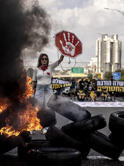 Demonstrantin in Tel Aviv (Israel) hält ein rotes Schild mit weißem Handabdruck in die Höhe; vor ihr liegt eine Barrikade aus brennenden Reifen, neben ihr steigt eine schwarze Rauchwolke in die Höhe (26.08.2025)
