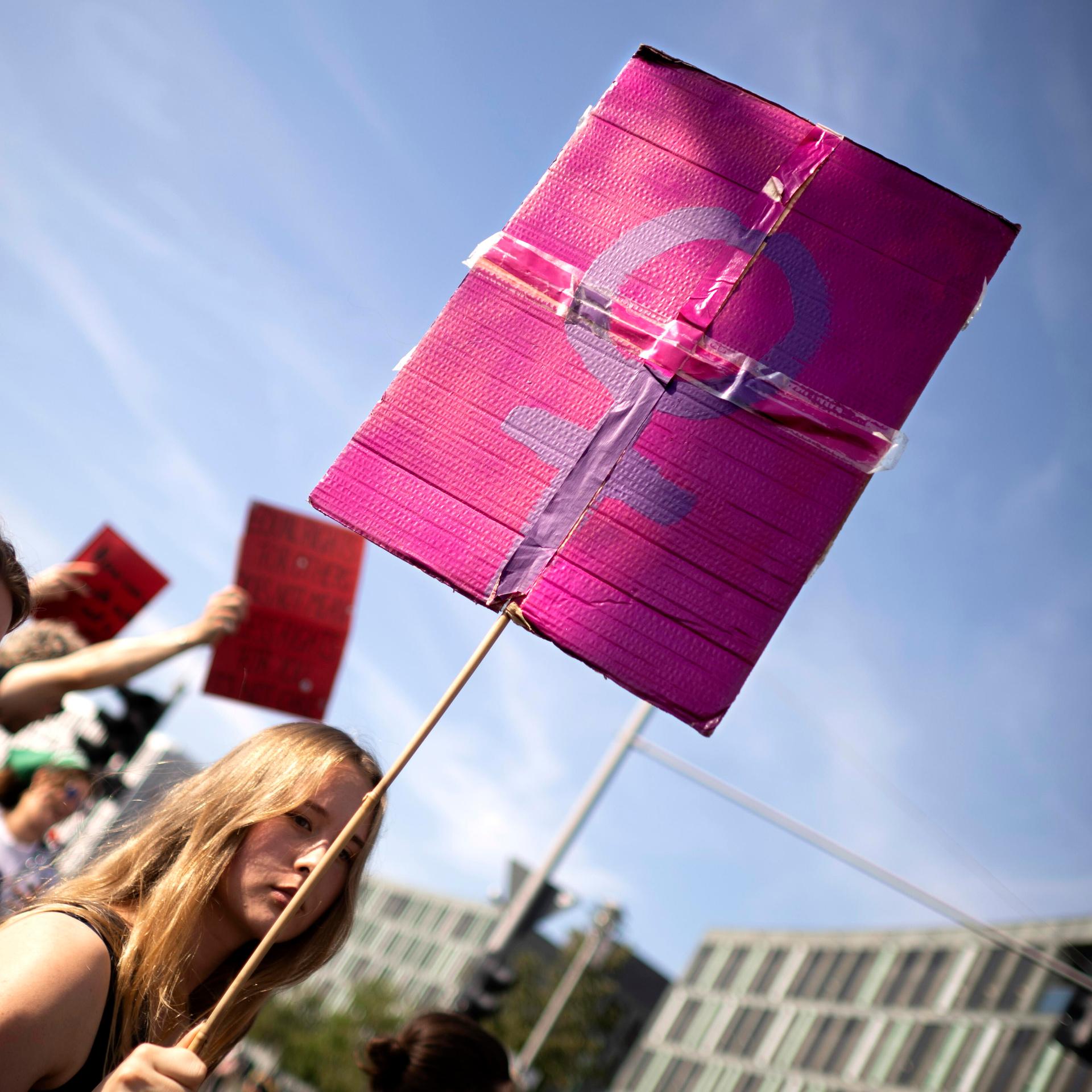 Demonstrantinnen in Berlin: Eine junge Frau trägt ein Schild, welches das weibliche Geschlechtssymbol, bestehend aus einem Kreis mit einem nach unten hängenden Kreuz, zeigt. Das lilafarbene Symbol hebt sich vom einem rosafarbenen Untergrund ab.