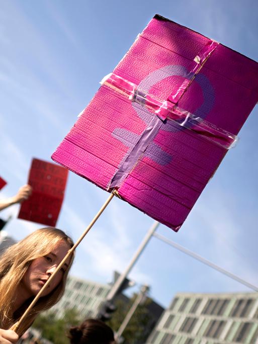 Demonstrantinnen in Berlin: Eine junge Frau trägt ein Schild, welches das weibliche Geschlechtssymbol, bestehend aus einem Kreis mit einem nach unten hängenden Kreuz, zeigt. Das lilafarbene Symbol hebt sich vom einem rosafarbenen Untergrund ab.