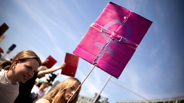 Demonstrantinnen in Berlin: Eine junge Frau trägt ein Schild, welches das weibliche Geschlechtssymbol, bestehend aus einem Kreis mit einem nach unten hängenden Kreuz, zeigt. Das lilafarbene Symbol hebt sich vom einem rosafarbenen Untergrund ab.