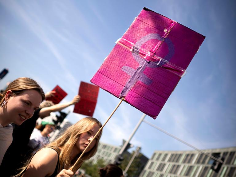 Demonstrantinnen in Berlin: Eine junge Frau trägt ein Schild, welches das weibliche Geschlechtssymbol, bestehend aus einem Kreis mit einem nach unten hängenden Kreuz, zeigt. Das lilafarbene Symbol hebt sich vom einem rosafarbenen Untergrund ab.