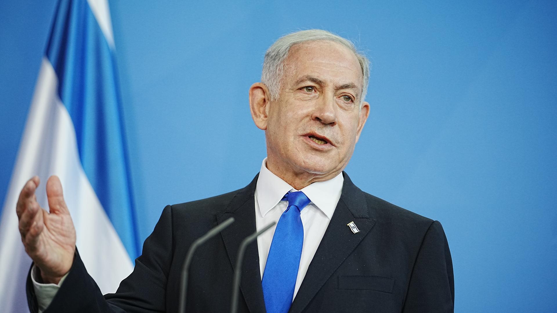 Benjamin Netanjahu steht vor einer israelischen Flagge und spricht. 