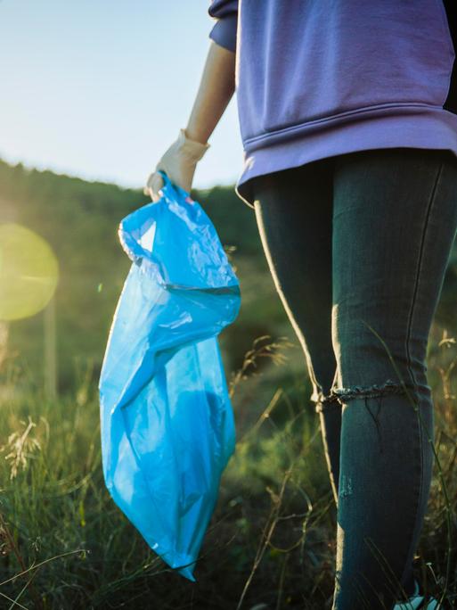 Eine ehrenamtliche Helferin läuft durch eine Wiese, in der einen Hand hält sie einen Müllsack und in der anderen ein Stück Plastik, das sie gerade aufgelesen hat. 