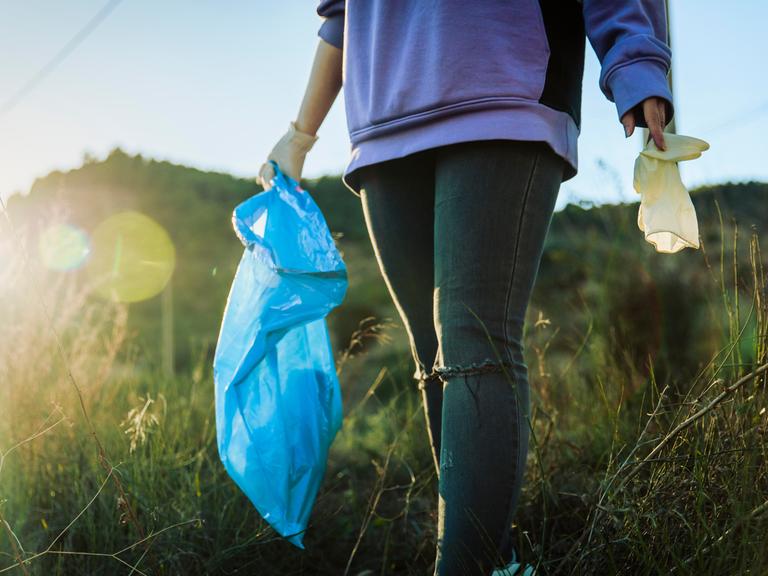 Eine ehrenamtliche Helferin läuft durch eine Wiese, in der einen Hand hält sie einen Müllsack und in der anderen ein Stück Plastik, das sie gerade aufgelesen hat. 