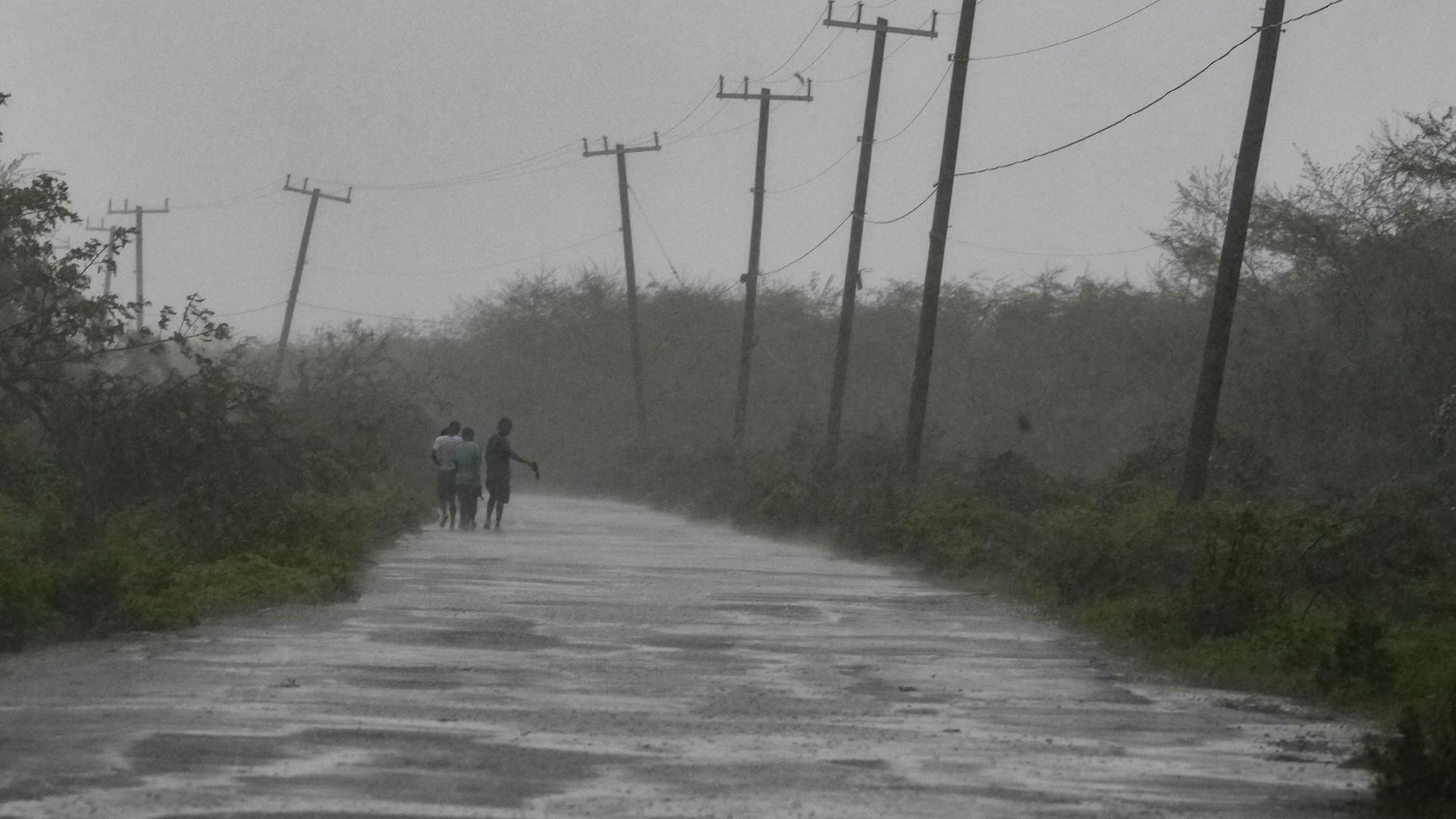 Blick auf eine überschwemmte Straße durch ein Naturgebiet mit Strommasten. Zwei Menschen gehen am Straßenrand durchs Wasser, man sieht sie aus der Ferne. Der Himmel ist grau; der Regen lässt das auch das gesamte Bild grau erscheinen. 