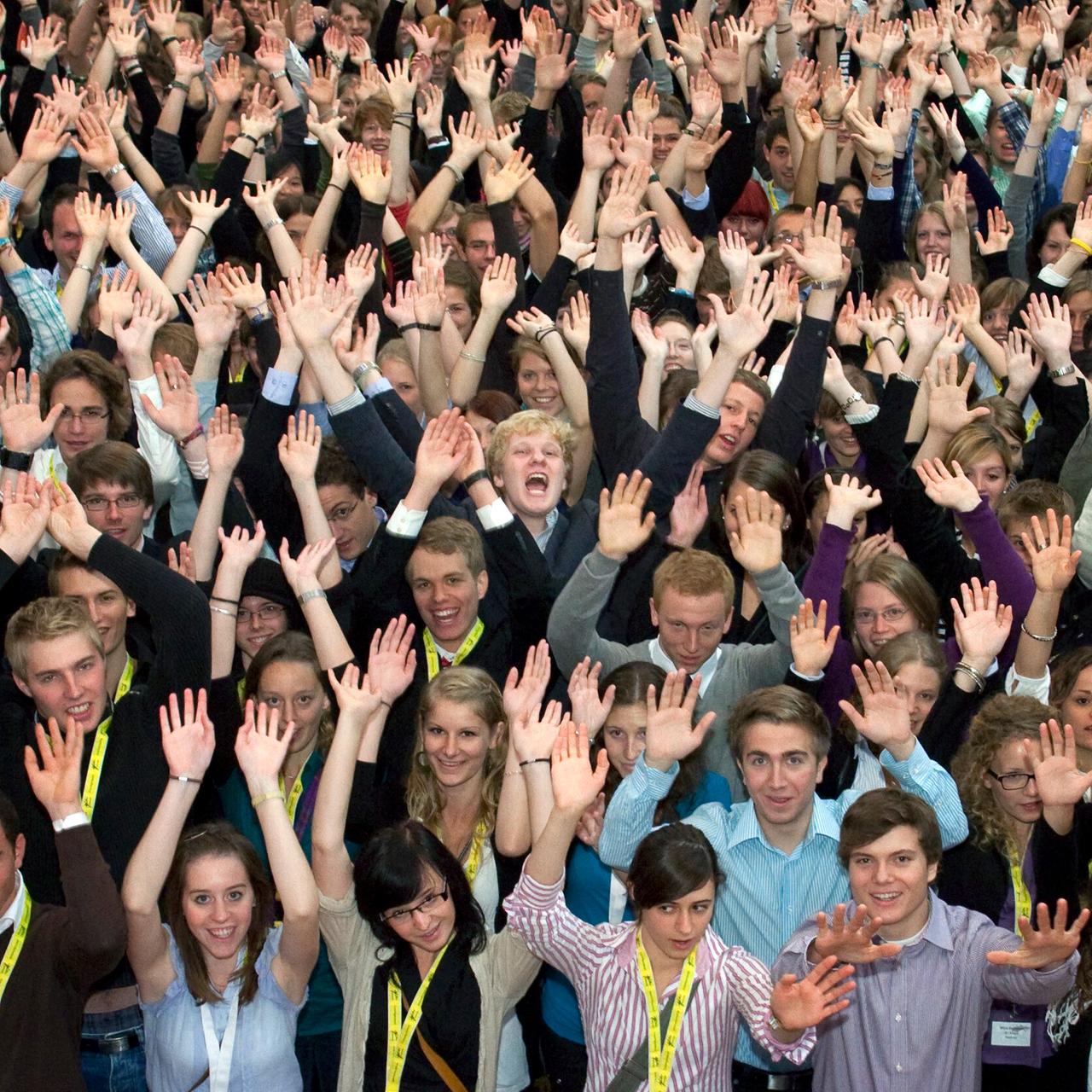 Eine eng beieinander stehende große Gruppe Jugendlicher reißt freudig johlend beide Arme hoch. 