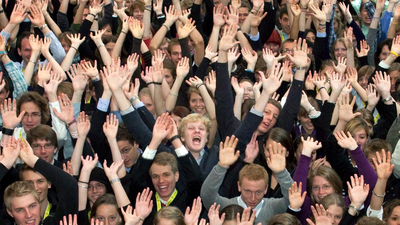 Eine eng beieinander stehende große Gruppe Jugendlicher reißt freudig johlend beide Arme hoch. 