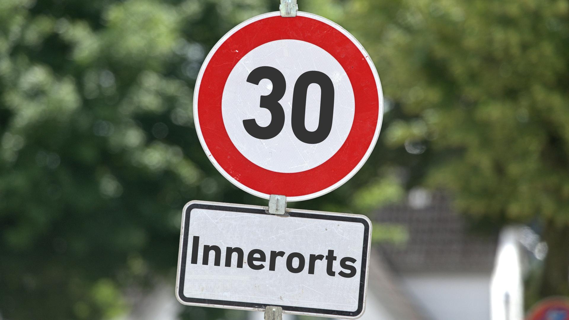 Verkehrszeichen in runder Form mit der Aufschrift "30" sowie ein Zusatzschild mit der Aufschrift: "Innerorts" an einem Schildermast. Im Hintergrund sieht man Bäume.
