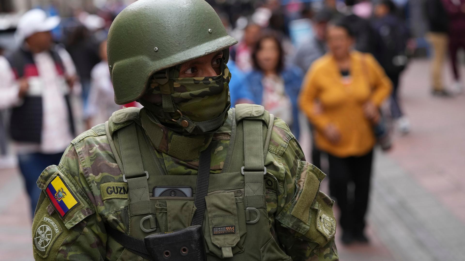 Ein Soldat mit vermummten Gesicht steht auf der Straße. Hinter ihm flanieren Passanten.