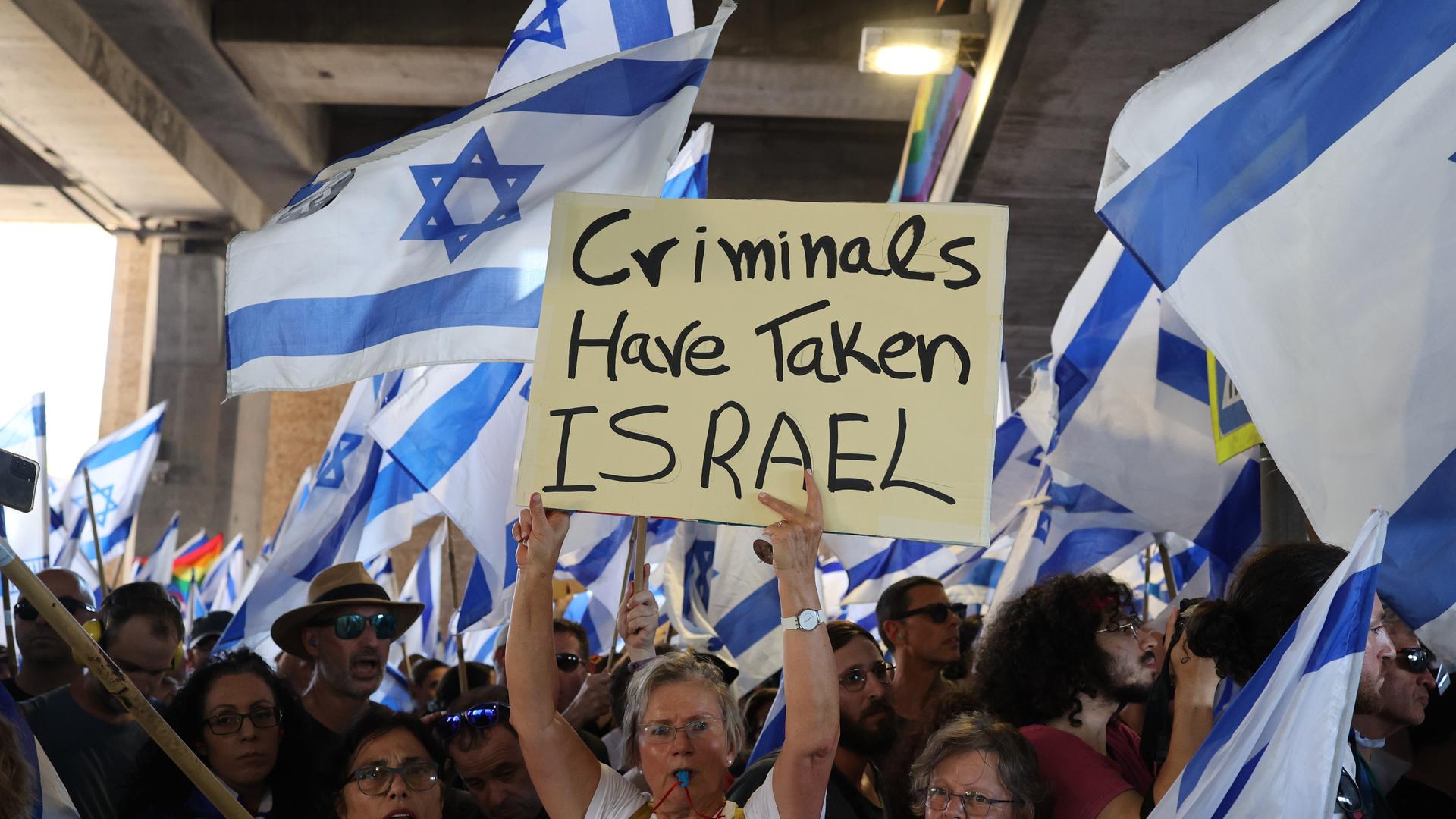 Israelische Demonstranten halten israelkische Fahnen und Plakate während einer Protestaktion. Auf einem Plakat ist auf englisch zu lesen: Kriminelle haben Israel übernommen.