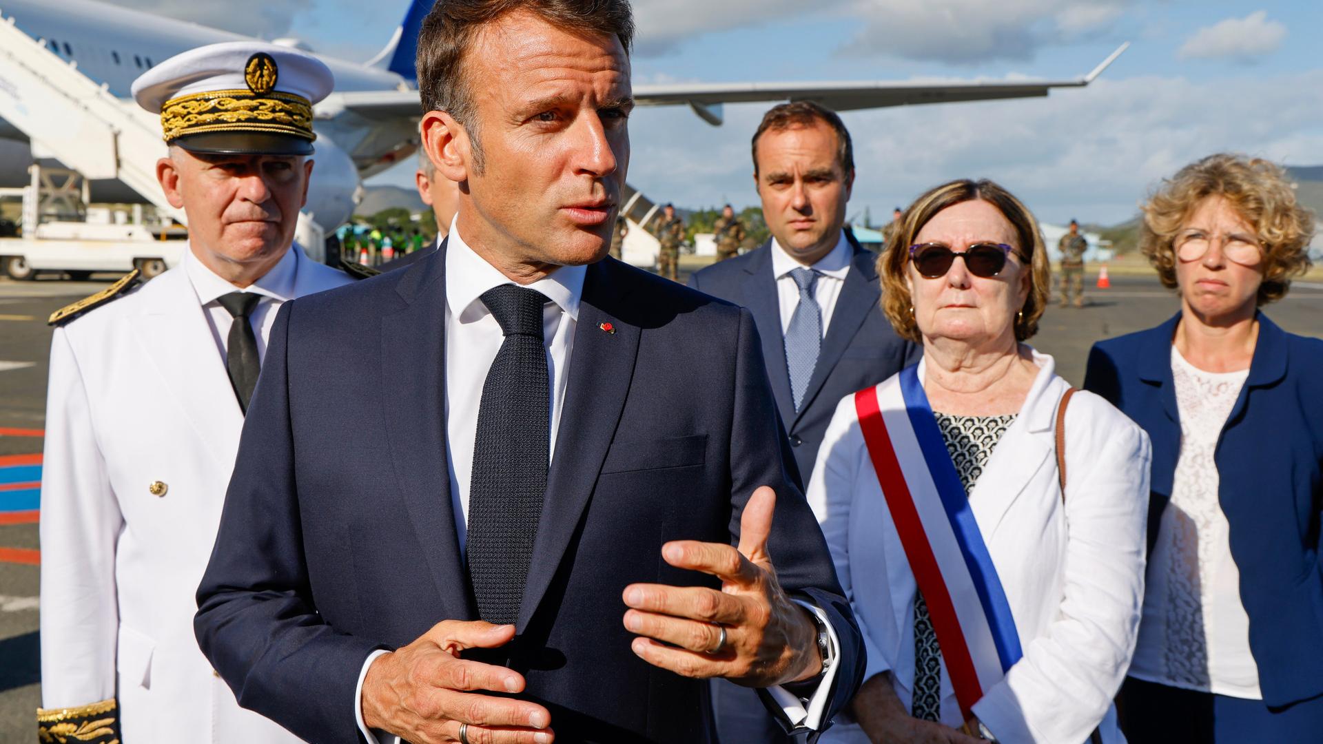 Frankreichs Präsident Macron steht auf einem Flugfeld, redet und gestikuliert. Um ihn herum stehen mehrere andere Menschen.