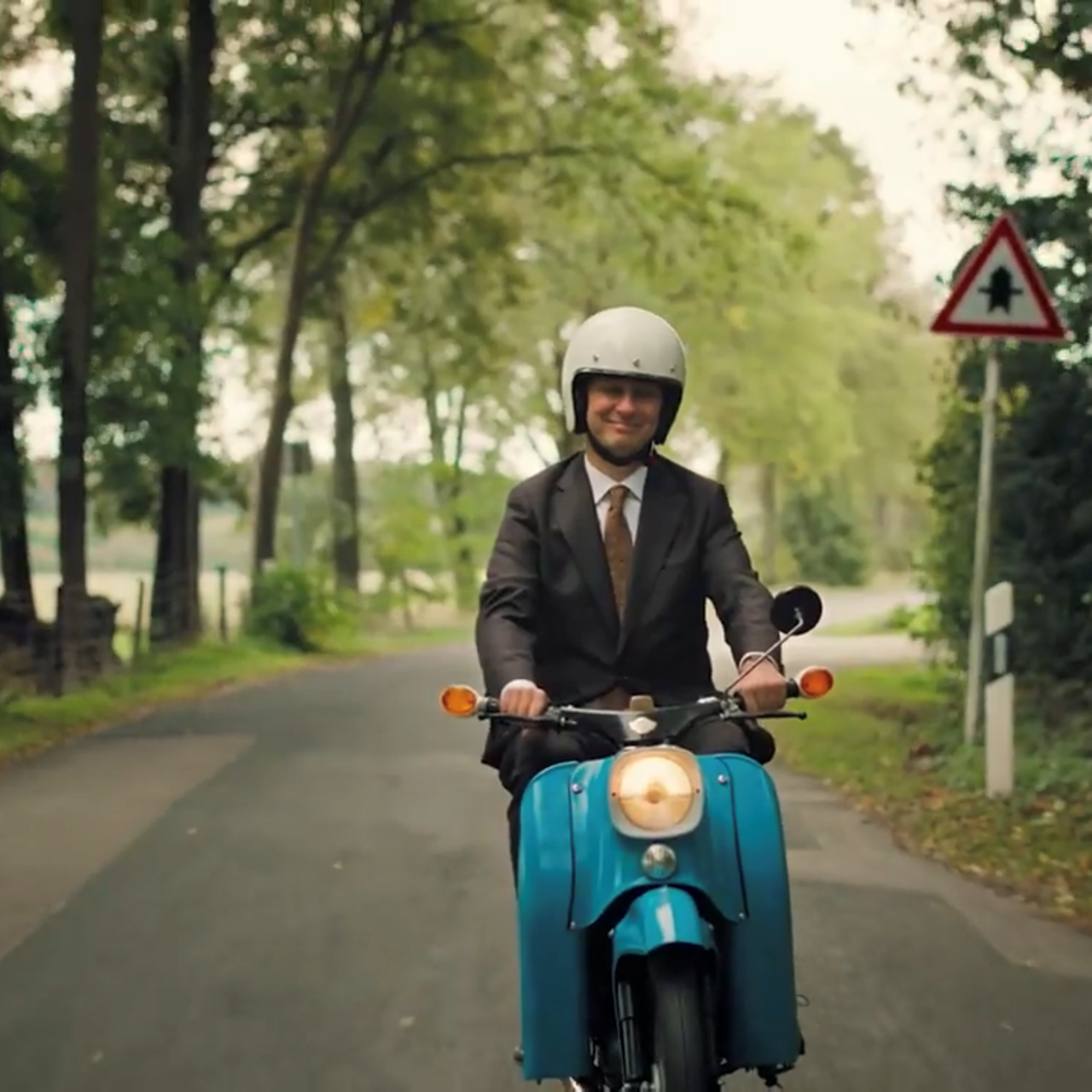 Jan Böhmermann fährt in braunem Anzug mit einem blauen Simson-Moped und weißem Helm über Land. 