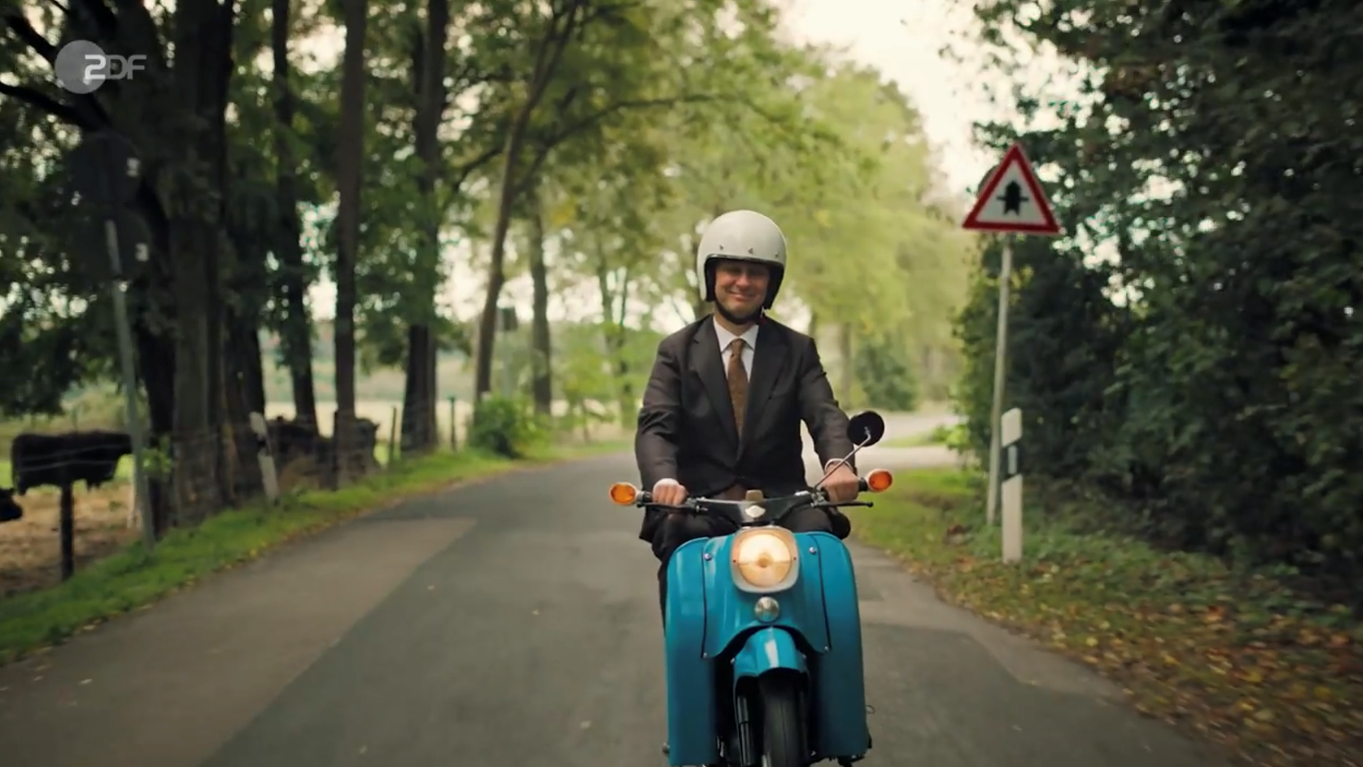 Jan Böhmermann fährt in braunem Anzug mit einem blauen Simson-Moped und weißem Helm über Land. 