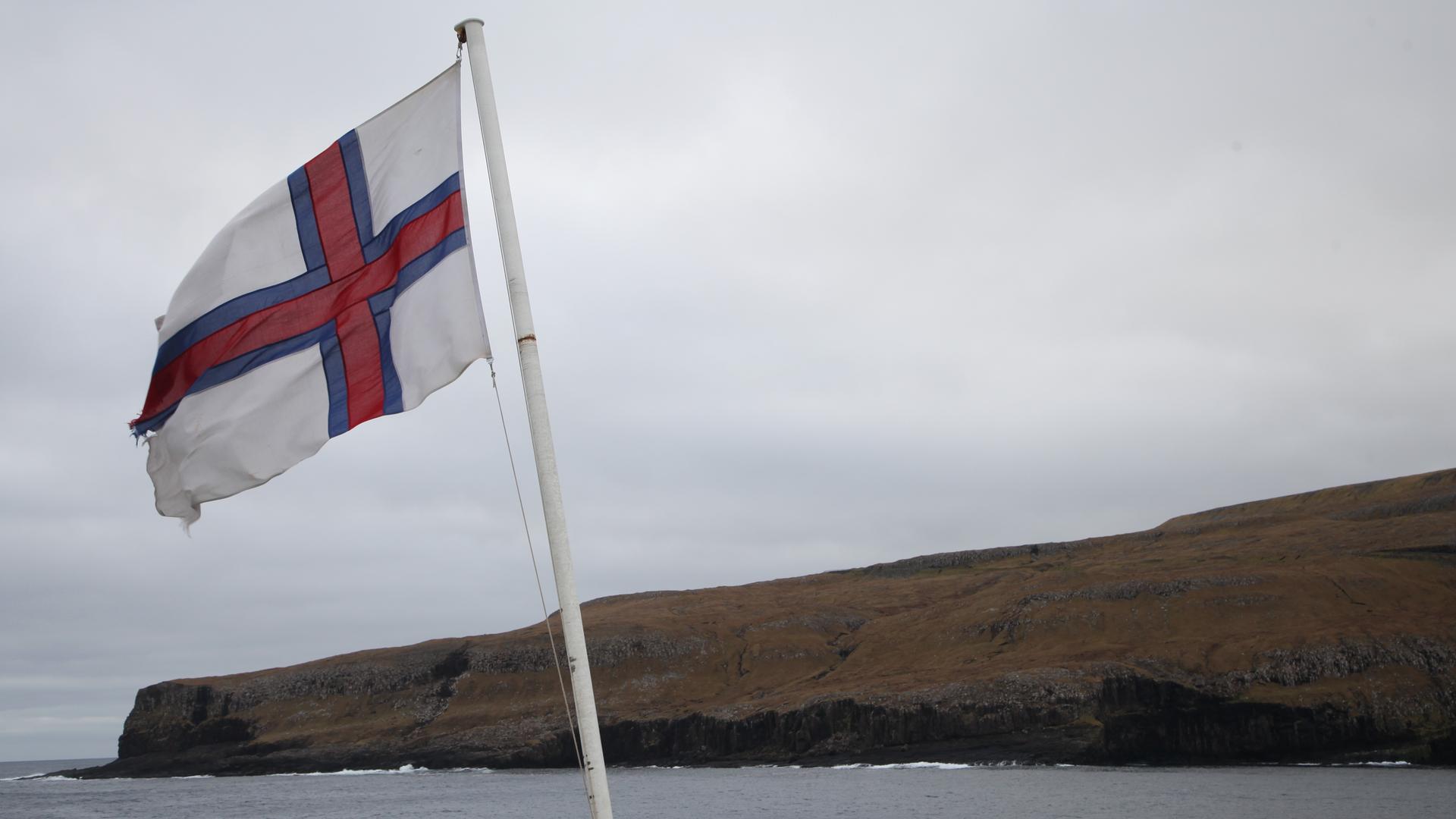 Eine Flagge der Färöer-Inseln weht an Bord einer Fähre bei der Einfahrt in den Hafen von Tvoroyri auf der färöischen Insel Suduroy im Wind. Eine Flagge der Färöer-Inseln weht an Bord einer Fähre bei der Einfahrt in den Hafen von Tvoroyri auf der färöischen Insel Suduroy im Wind.